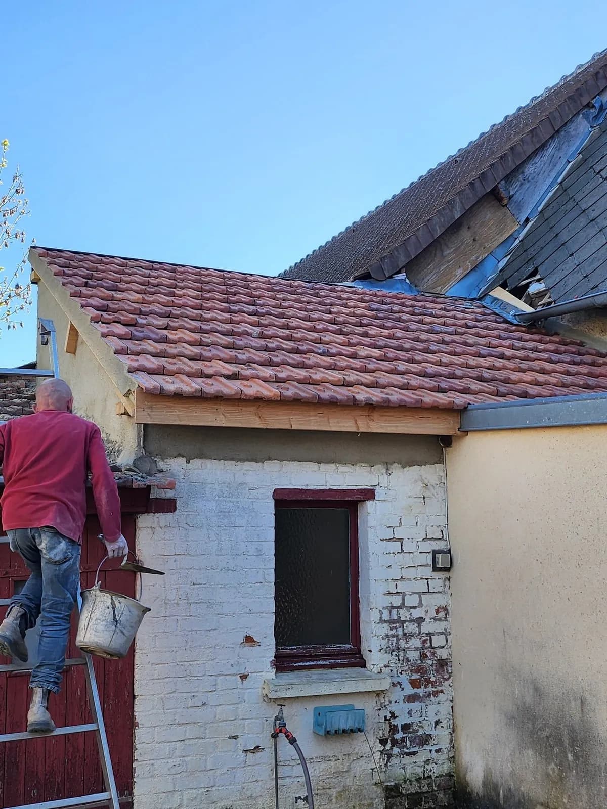 Couvreur de Bouthors Couverture sur l’échelle, seau à la main, au bord de la toiture neuve d’une annexe en tuiles à emboîtement