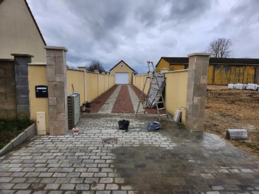 Cour pavée entre deux murs enduits, piliers en pierre et allée vers le garage, chantier de SJ Rénovation, partenaire de Bouthors Couverture