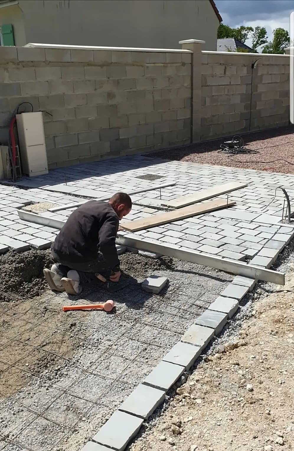 Ouvrier de Bouthors Couverture à genoux posant des pavés gris dans une cour, règle et maillet à côté, mur de clôture neuf au fond